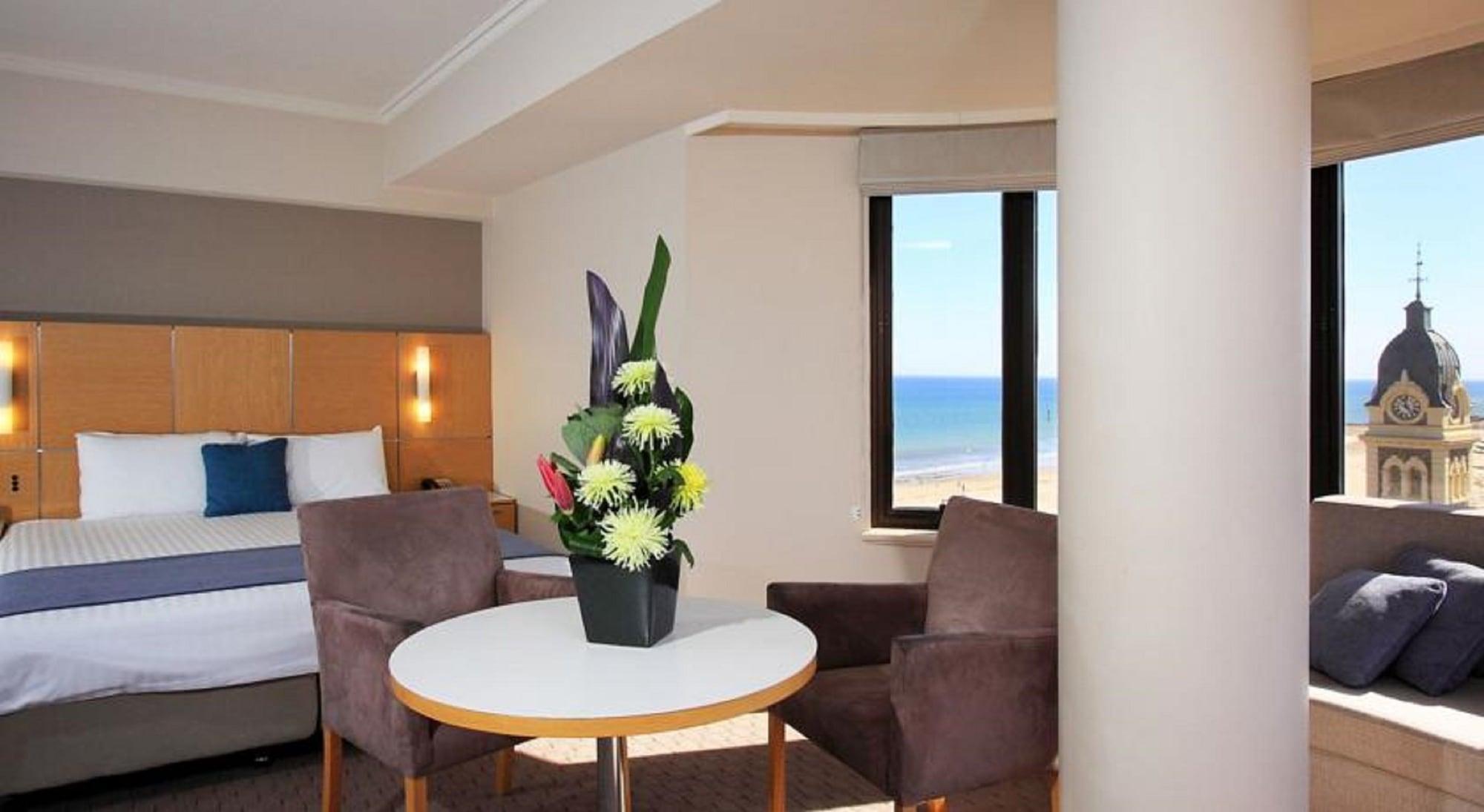 Stamford Grand Adelaide Hotell Glenelg Exteriör bild The photo shows a cozy hotel room with a comfortable seating arrangement. There are two brown chairs positioned around a round table, which features a decorative flower arrangement in a black vase. The background includes a window that offers a view 
