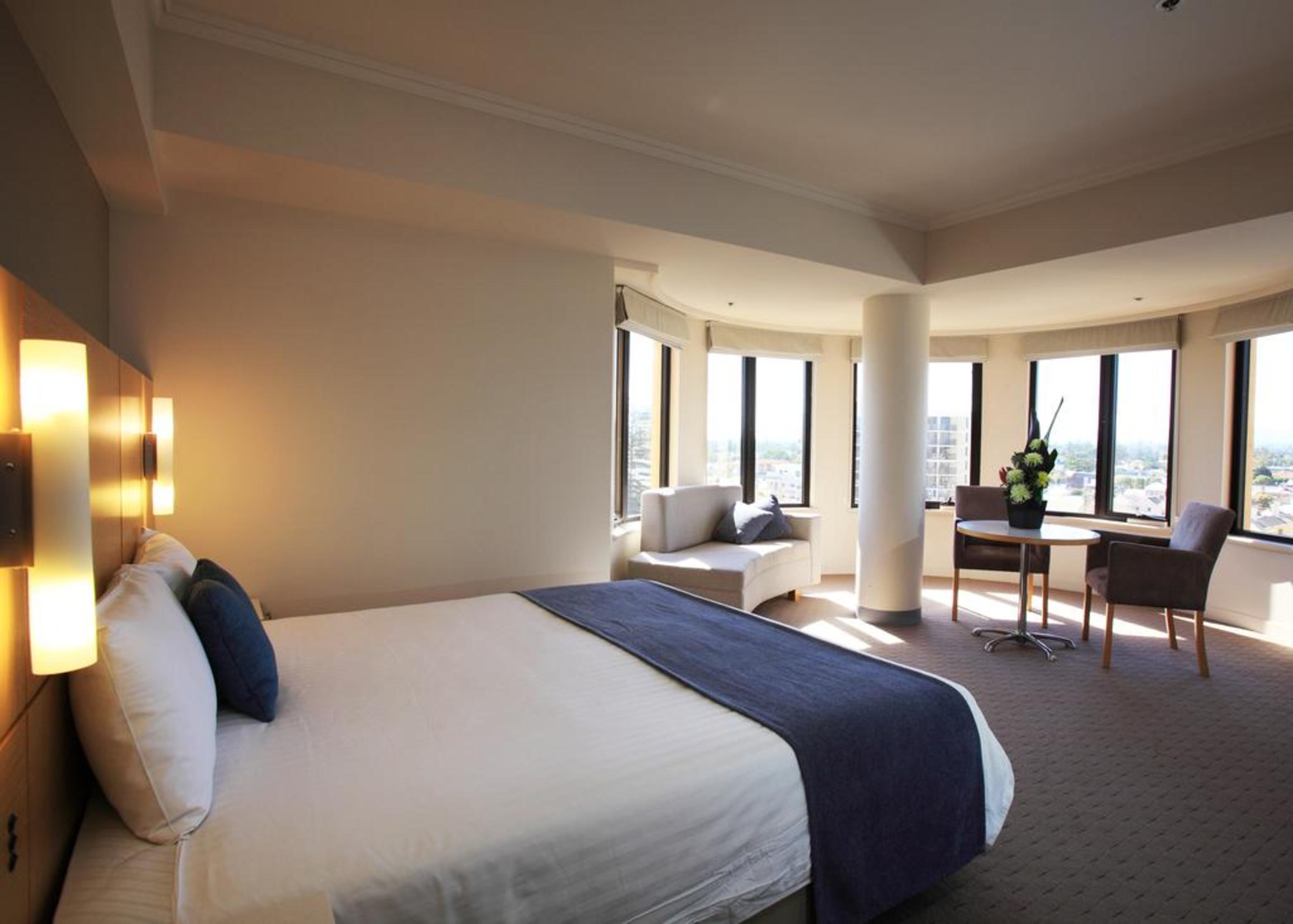 Stamford Grand Adelaide Hotell Glenelg Exteriör bild The photo shows a hotel room with a modern and spacious design. There is a large bed with white linens and a dark blue throw blanket. The room features multiple windows that allow plenty of natural light, and there is a small seating area near the wi