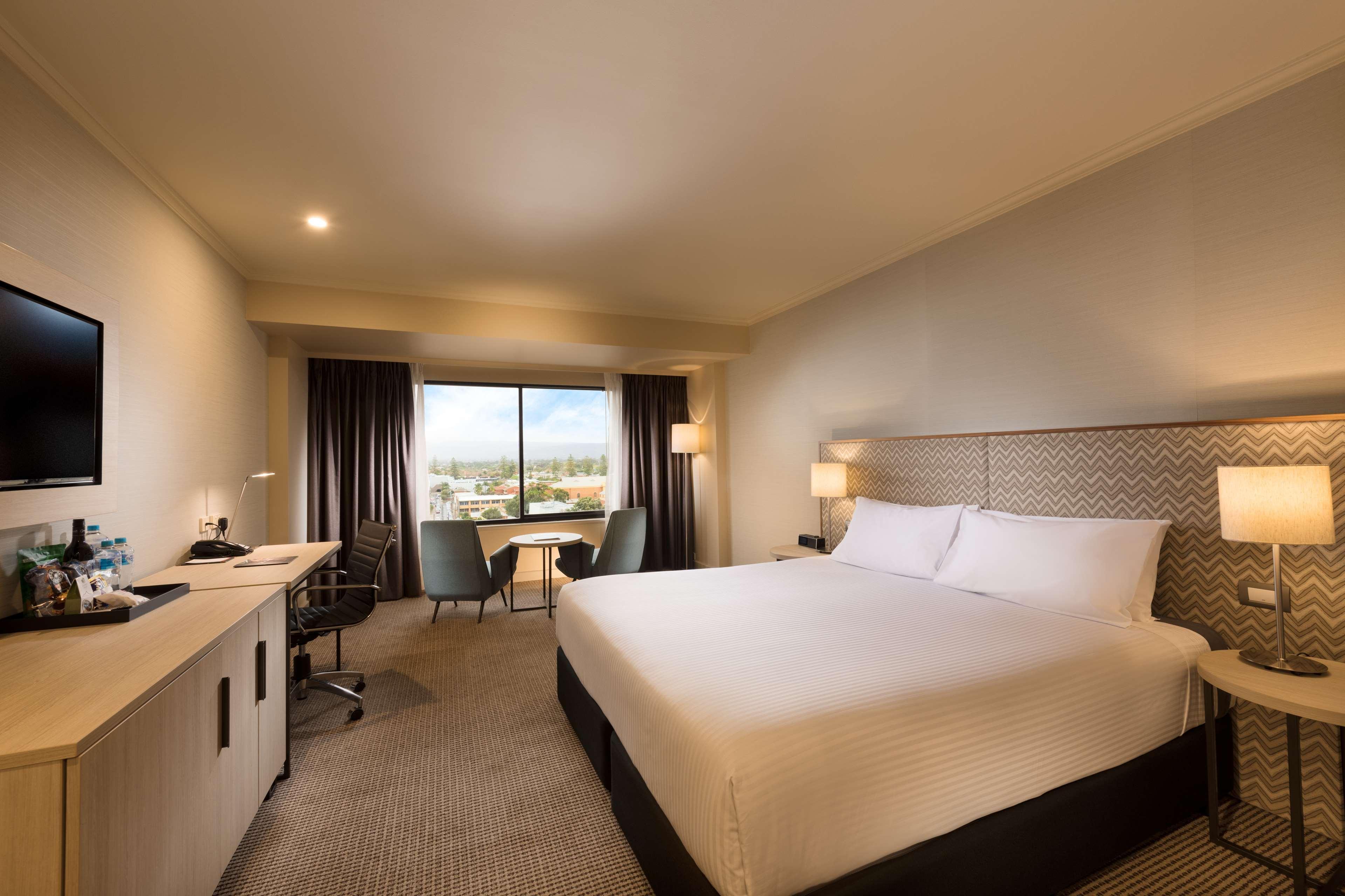 Stamford Grand Adelaide Hotell Glenelg Exteriör bild The photo shows a modern hotel room. It features a large bed with white linens, a desk with a chair, and a flat-screen TV mounted on the wall. There are two armchairs and a small table near a large window that lets in natural light, providing a view 