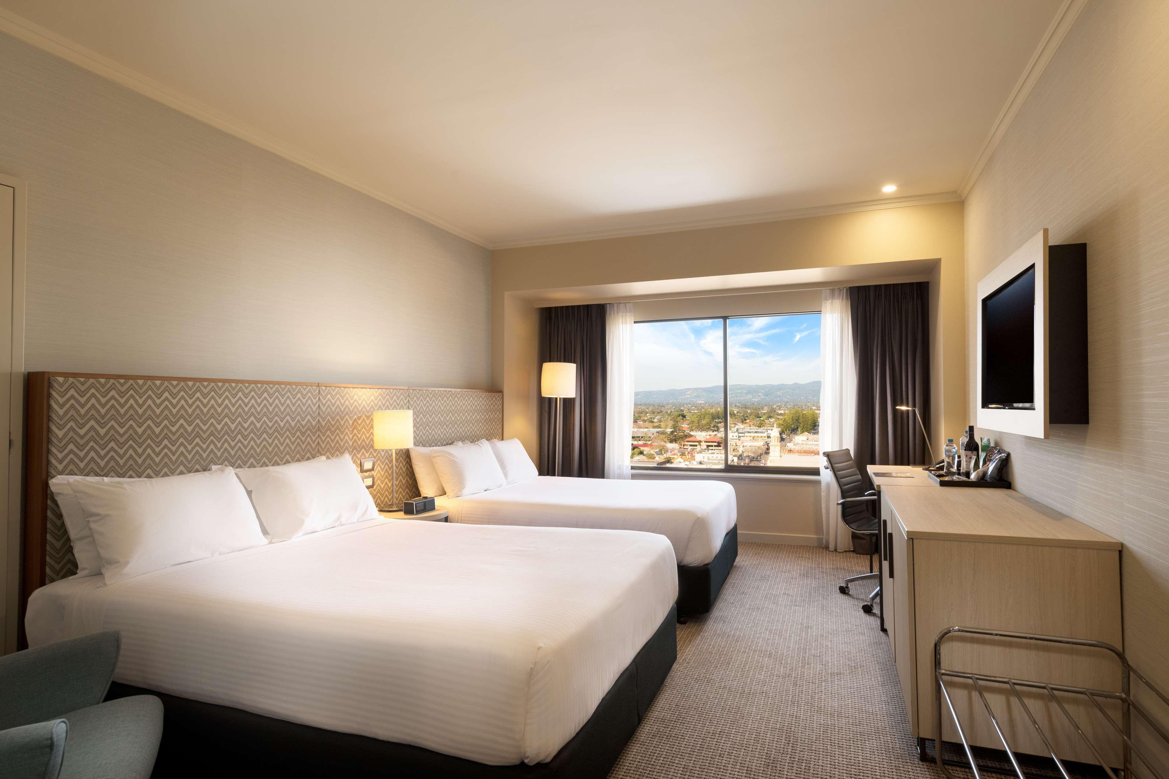 Stamford Grand Adelaide Hotell Glenelg Exteriör bild The photo shows a modern hotel room featuring two beds with white linens. There are bedside tables with lamps next to each bed. A large window provides natural light and offers a view of the outside, with curtains on either side. The room has a neutr
