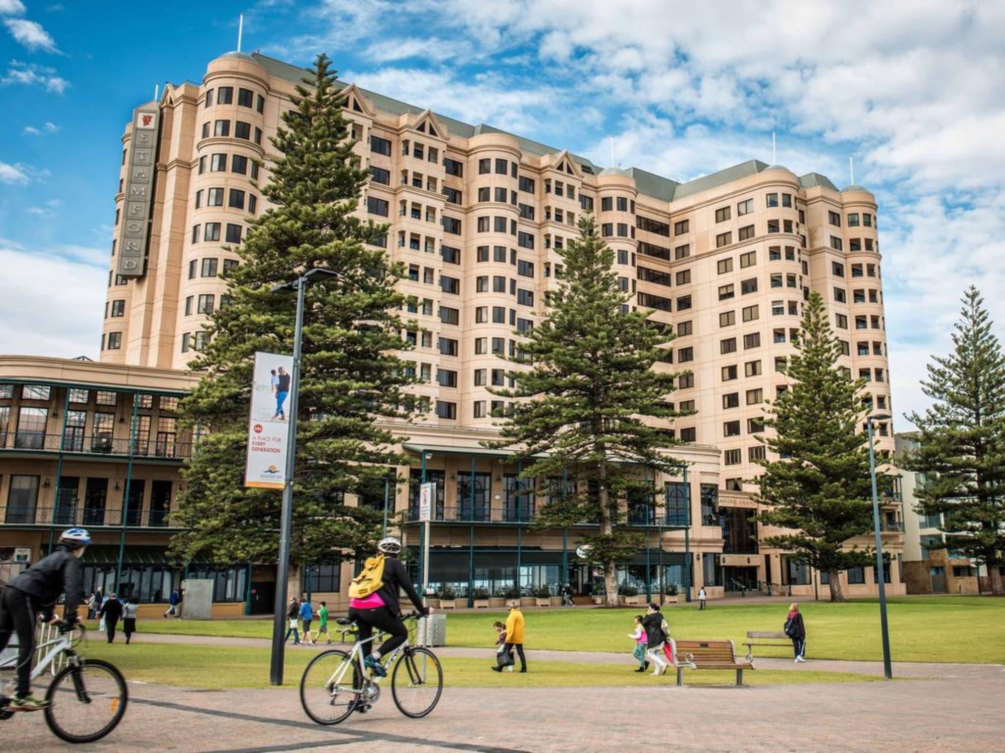 Stamford Grand Adelaide Hotell Glenelg Exteriör bild The photo depicts a modern building with a large facade, likely a hotel or commercial establishment, situated near a spacious green area. Surrounding the building are several tall trees, providing shade and enhancing the aesthetic appeal. In the fore