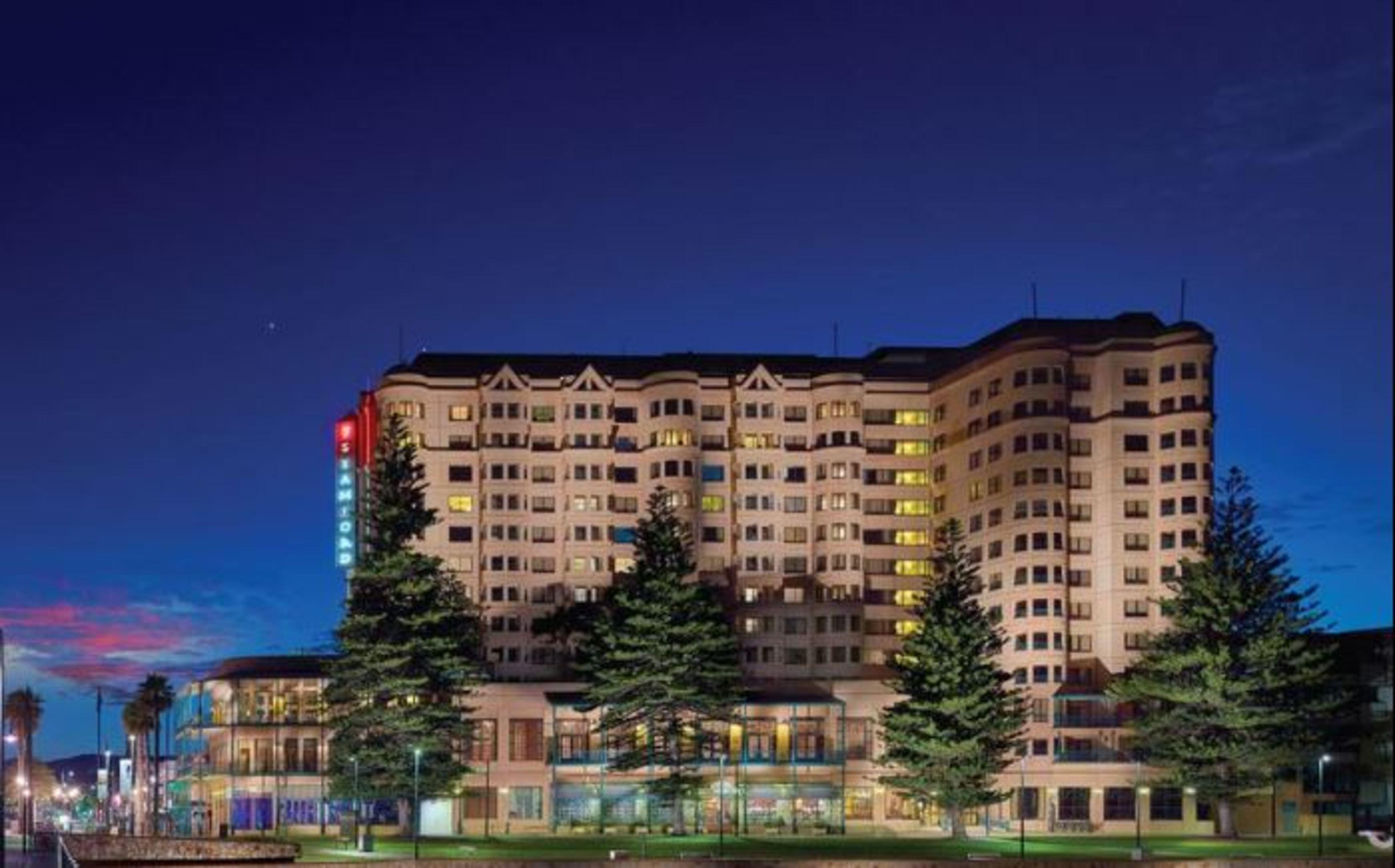 Stamford Grand Adelaide Hotell Glenelg Exteriör bild The photo displays a multi-story hotel building at dusk, illuminated by various lights. The architecture features a mix of modern and traditional design elements, with numerous windows showcasing a warm glow. Surrounding the hotel are several tall tr