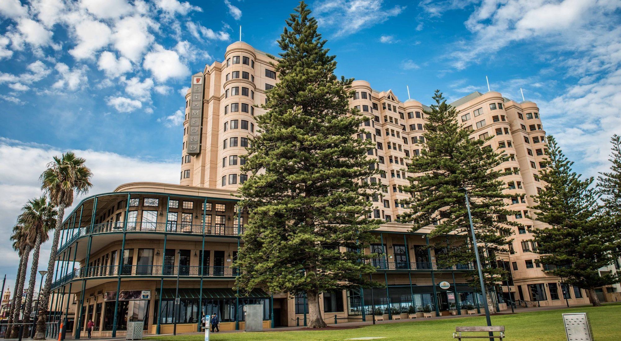 Stamford Grand Adelaide Hotell Glenelg Exteriör bild The image shows a large building with a distinctive architectural style, characterized by a tan or beige color. The structure appears to have multiple floors and rounded edges. In front of the building, there are several tall green trees, which add t